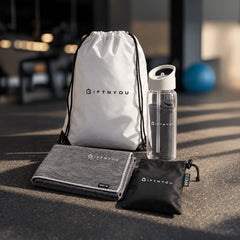 The Active Refresh Kit by GiftNYou with clear bottle with carry handle and grey towel inside, additional grey sports towel, black drawstring storage pouch in a fitness setting